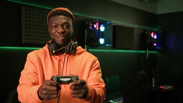 Cheerful African American Male Gamer Dressed in an Orange Outfit with Big Black Headphones Around