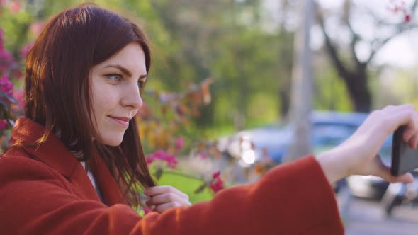 Young Attractive Redhaired Woman Makes a Selfie on the Background of Spring Flowers of Cherry or