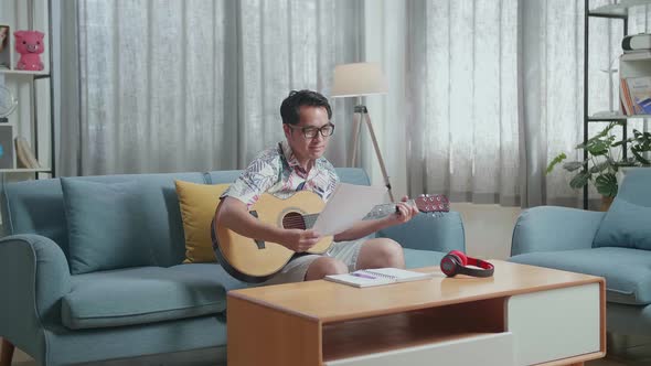 Asian Man Composer With Note Book And Looking At Paper While Playing Guitar At Home