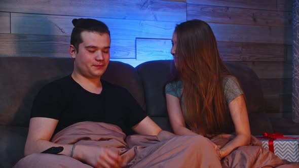 Young Cute Couple Sitting on a Couch Under a Blanket  the Woman Gives Her Boyfriend a Christmas Gift
