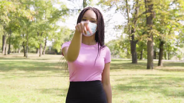 A Young Black Woman in a Face Mask Points at the Camera and Nods in a Park on a Sunny Day