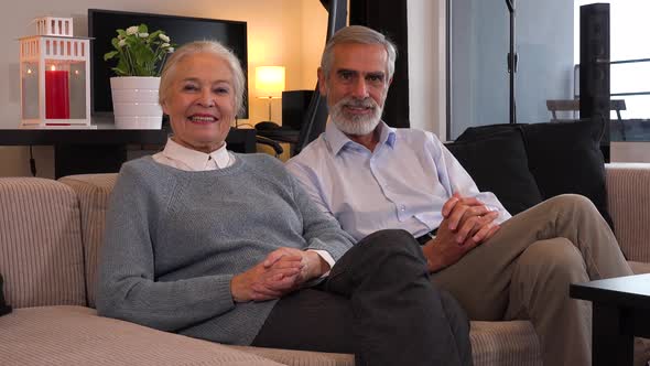 An Elderly Couple Sits on A Couch in An Apartment and Smiles at The Camera