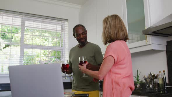 Diverse senior couple in kitchen using laptop and drinking wine