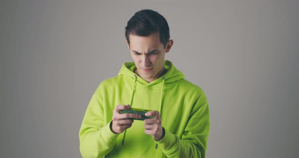 Handsome Young Man in Green Sweatshot Playing Game on Smartphone at Grey Studio Wall