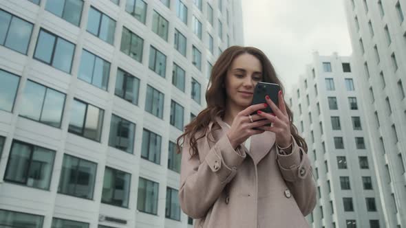 Cheerful young Caucasian woman standing in city street and texting on mobile phone with smile.