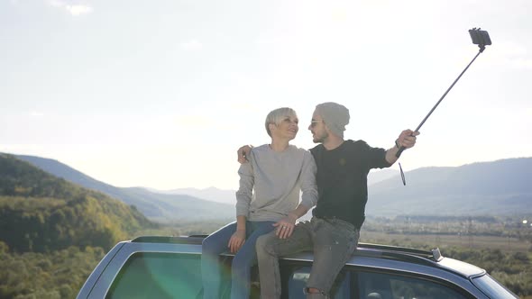 Happy Couple Sitting on Roof of Car and Possing on the Smart Phone Camera in the Mountain