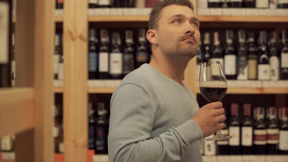 Handsome Man Standing with Glass of Red Wine in Liquor Store. Woman in the Background