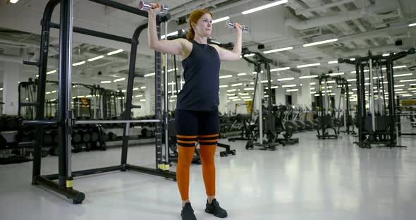 Athletic Girl in the Gym Doing Standing Exercises with Dumbbells on Her Arms and Shoulders