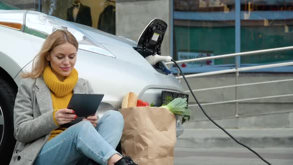 Woman Use Tablet and Leans on Electric Car That Is Charging. Plug Charging an Electrical Car