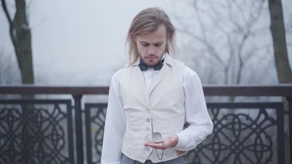 Stylish Caucasian Guy with Long Hair and Beard Standing at Bridge and Checking Time on Vintage Hand