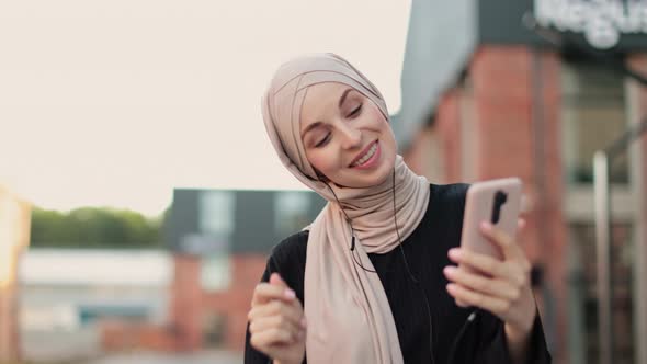 Joyful Muslim Girl in Hijab Wearing Wireless Headphones Listening Songs Online on Smartphone