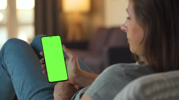 Woman at Home Lying on a Sofa and Using Smartphone with Green Mock-up Screen in Vertical Mode. Girl