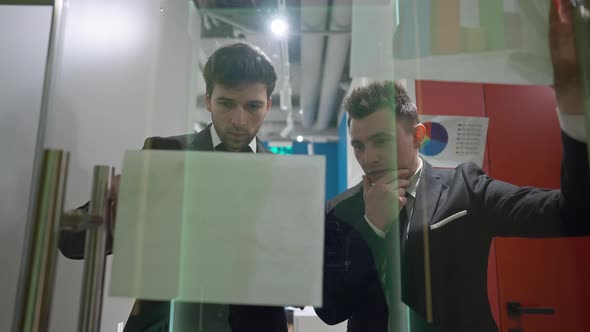 Two Thoughtful Men Analyzing Strategy Looking at Paperwork Standing Behind Glass Door in Office