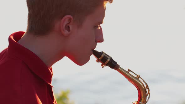 A young guy plays the saxophone near the river bank at sunset. Close-up.