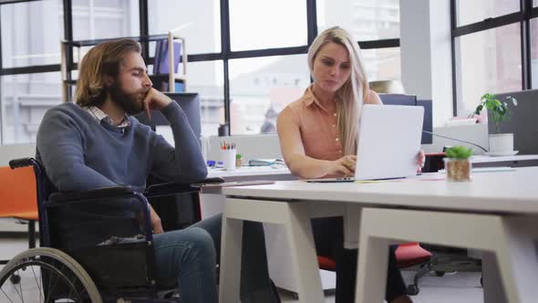 Disabled caucasian businessman sitting on wheelchair using laptop talking to coworker in office