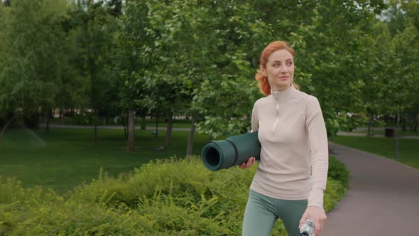 Confident woman walking with rolled yoga mat outdoor, morning gym routine.