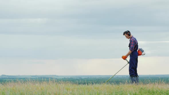 Young Man Mowing Grass with a Trimmer on a Picturesque Meadow