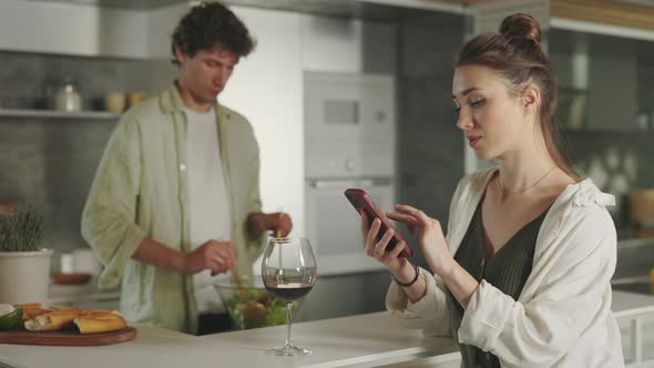 Man Cooking Salad While Woman Using Smartphone