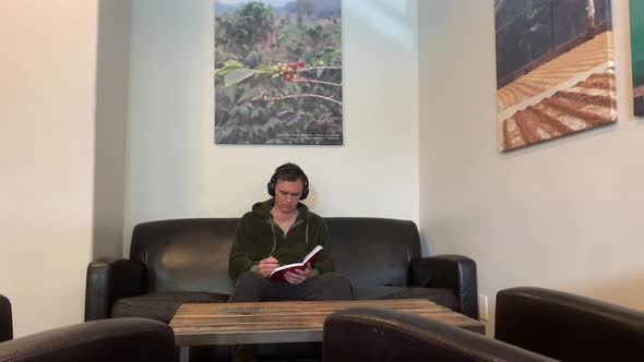 A man sits on a sofa with headphones and reads and writes in a book