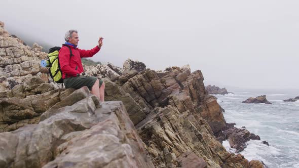 Senior hiker man with backpack sitting on the rocks and taking pictures on smartphone while hiking