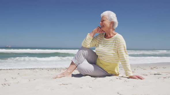 Happy senior caucasian woman with hand on chin sitting on the beach enjoying the view