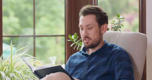 Close Up Portrait of Young Bearded Man Reading Book While Sitting in White Chair Young Man Relaxing