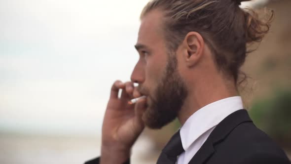 Confident Man in Business Suit with Tied Hair Thoughtfully Watching in Distance at Sea and Smoking