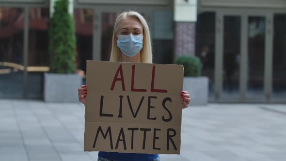 Young Woman Blonde in Medical Mask Stands with a Cardboard Poster ALL LIVES MATTER in a Public Place