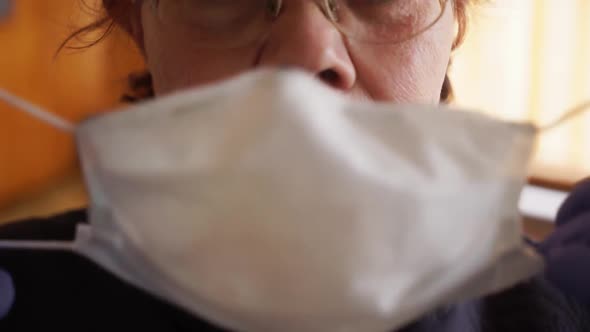 An Elderly Woman Puts on a Protective Medical Mask Near the Window, Looks at the Camera. COVID-19