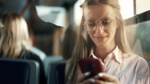 A Smiling Young Woman is Using Smartphone Social Media Reading Messages in a Bus and Smiling