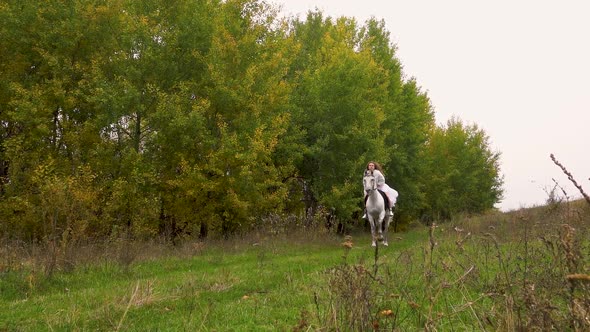 Long-haired Woman in White Dress Is Riding Horse Along Wood