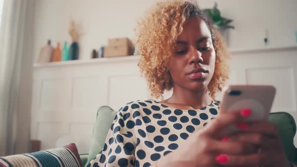 Young African American Woman Chatting in Phone Sits on Sofa in Living Room