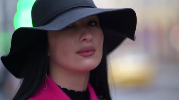 Portrait of a Brunette in a Black Hat and a Pink Coat Against a Background of Blurred Lights