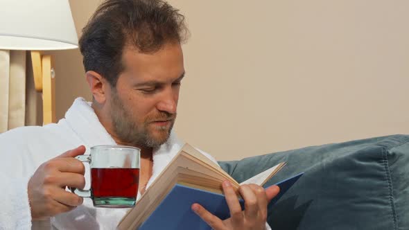 Mature Man Having Aromatic Hot Tea, Reading a Book at Home, Wearing Bathrobe 1080p