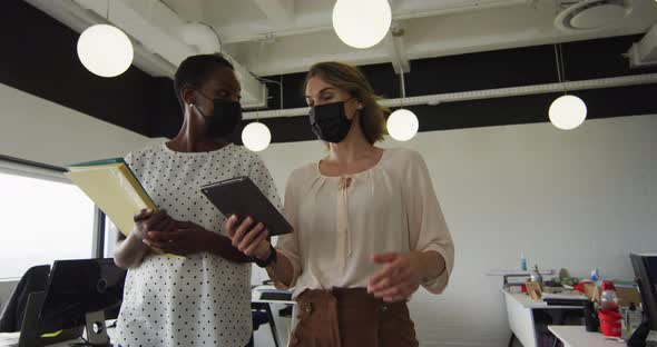 Two diverse female colleagues wearing face masks, walking, and discussing in office