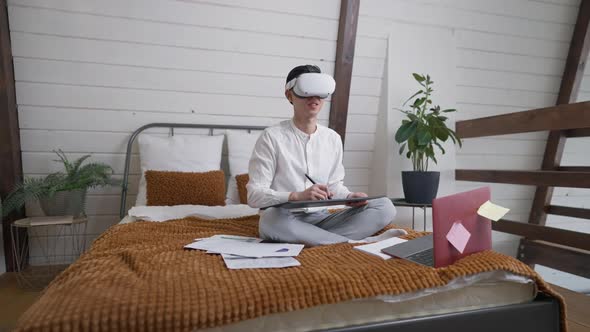 Wide Shot of Positive Creative Young Asian Man in VR Headset Sitting on Bed Drawing on Graphics