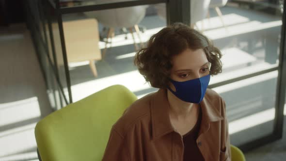 A Young Attractive Girlstudent is Sitting in a Blue Protective Face Mask in the Hall and Looking at