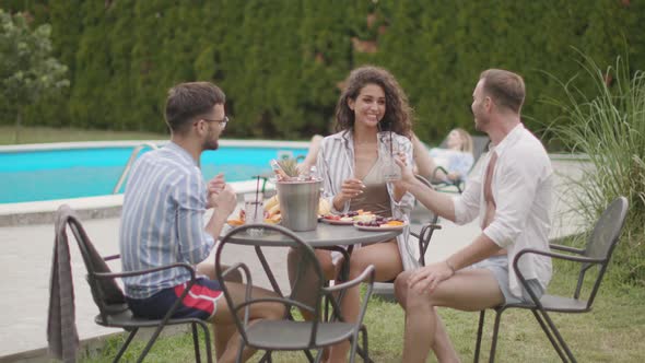 Group of happy young people cheering with drinks and eating fruits by the pool in the garden