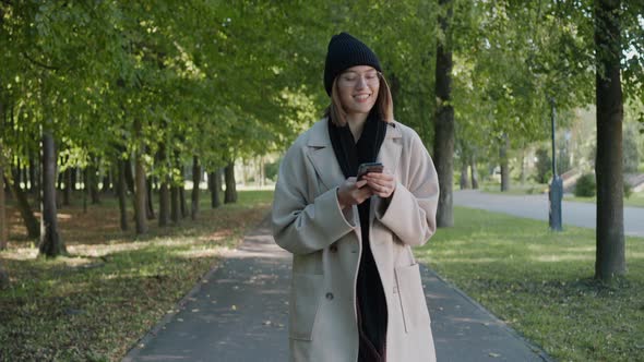 Young Girl Walks in the City Park on a Sunny Autumn Day Texting and Smiling