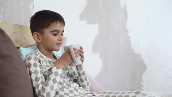 Little Boy Drinks Tea From Mug Sitting on Bed