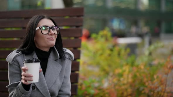 Funny Woman is Drinking Hot Tea or Coffee From Cardboard Cup in Park in Autumn Sitting on Bench