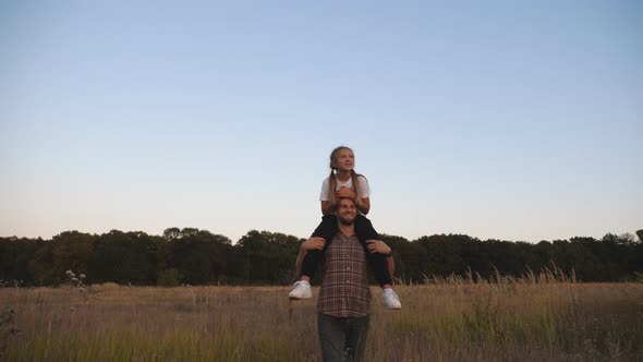 Young Father Walks with Cute Small Daughter Sitting on His Shoulders Through Meadow