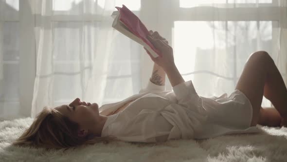 Portrait of Young Caucasian Girl Lying on Bed and Reading Book. Pretty Blond Woman in White Shirt