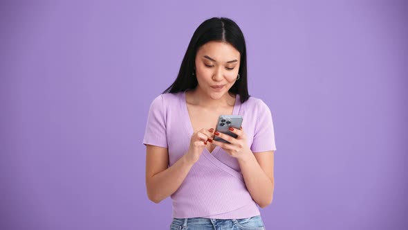 Cheerful Asian brunette woman wearing purple t-shirt texting by mobile