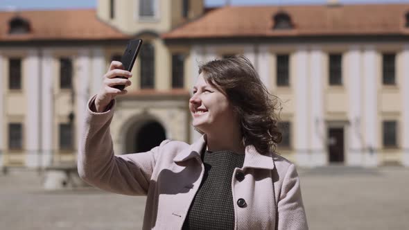 Happy Woman in Coat Takes Photos on Smartphone in Yard of Medieval Castle