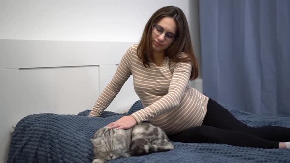 A Young Pregnant Woman with Glasses is Stroking a Cat