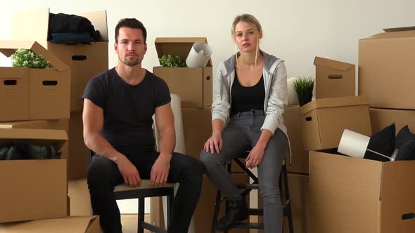 A Moving Couple Sits on Chairs and Looks Seriously at the Camera in an Empty Apartment