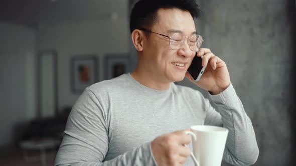 Asian smiling man wearing eyeglasses talking on mobile and drinking coffee