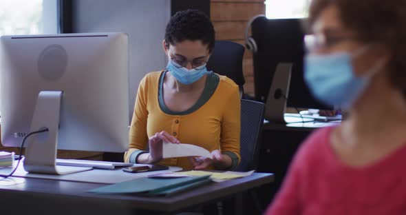 Caucasian woman wearing face mask reading documents and using computer at modern office