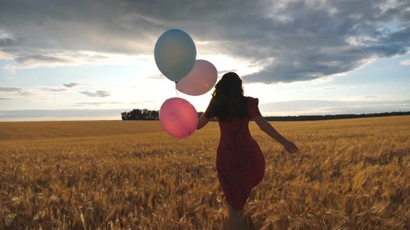 Rear View of Happy Girl in Red Dress Running Through Golden Wheat Field with Balloons in Hand at